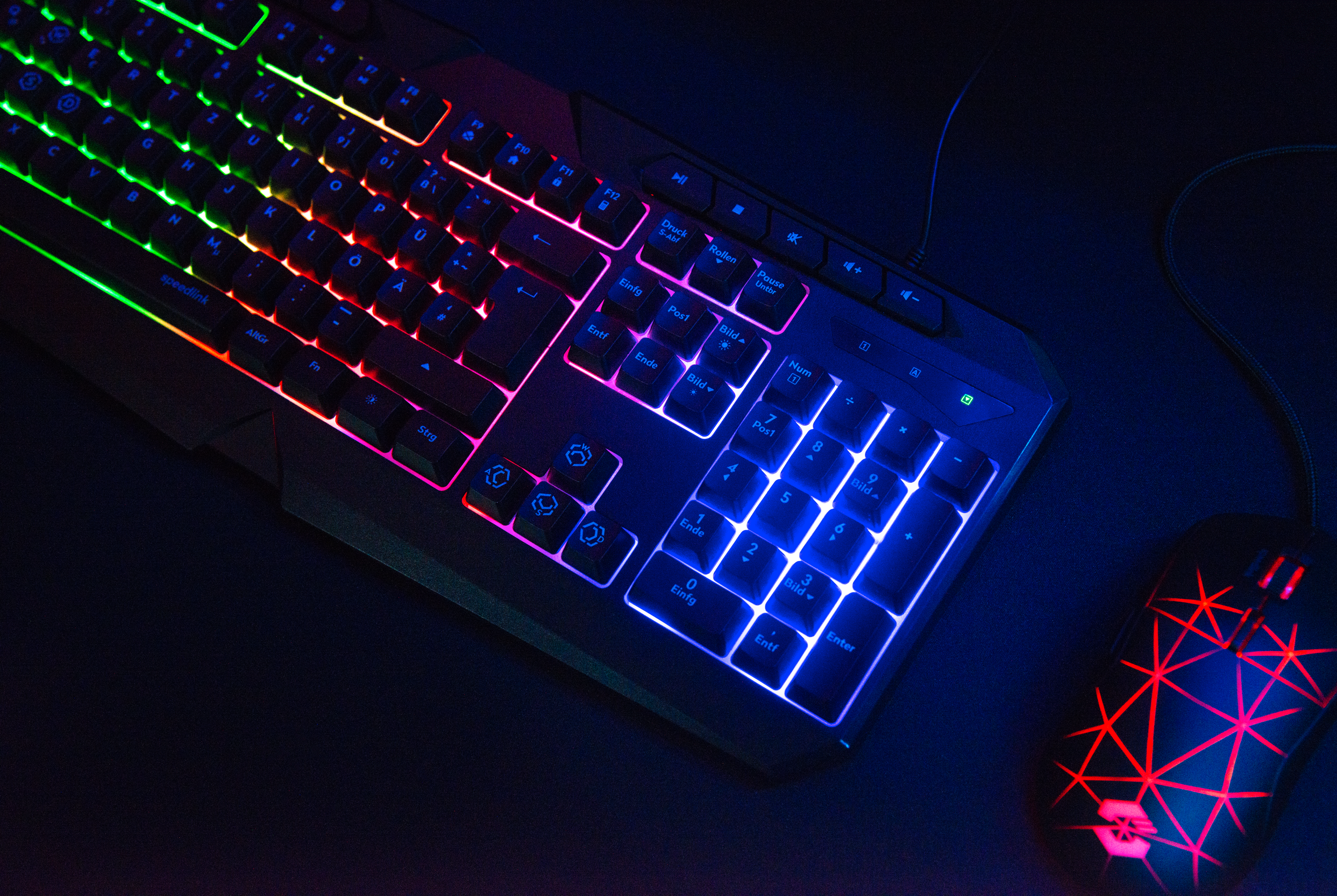 Beleuchtete Gaming-Tastatur mit RGB-Farben und einer schwarzen Gaming-Maus mit roten LED-Linienmuster auf einem dunklen Hintergrund