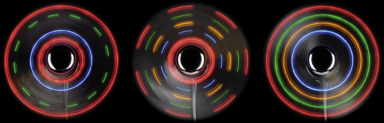 Drei rotierende LED-Fidget Spinner mit leuchtenden, bunten Lichtspuren auf schwarzem Hintergrund