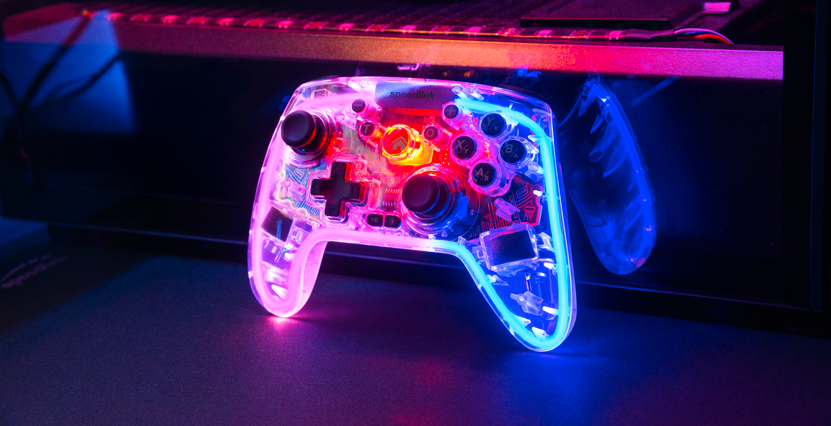 Transparenter Gaming-Controller mit bunter LED-Beleuchtung vor einem Computer in neonblauer und pinker Atmosphäre