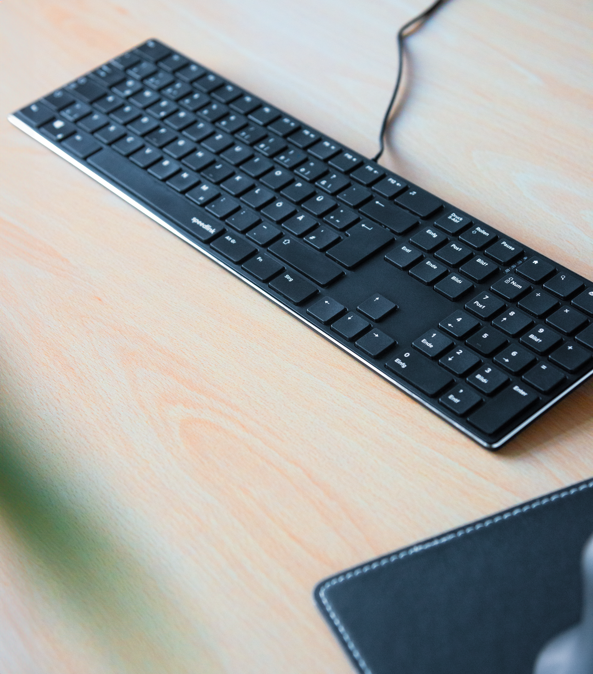 Flache schwarze Computer-Tastatur mit Kabel auf einem hellen Holzschreibtisch, neben einem Mousepad