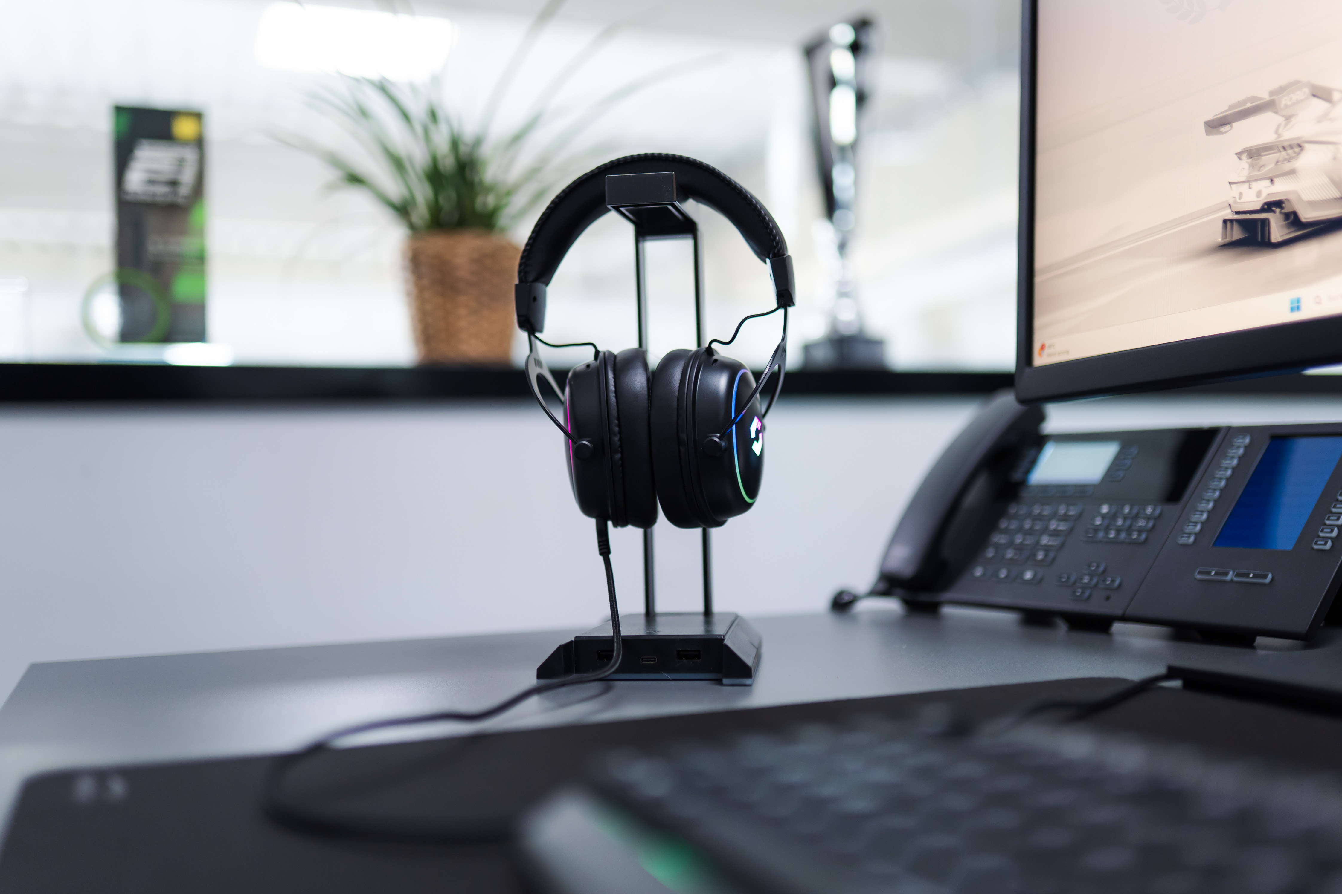 Ein schwarzes Headset mit LED-RGB-Beleuchtung steht auf einem Ständer neben einem Monitor und Telefon auf einem Schreibtisch