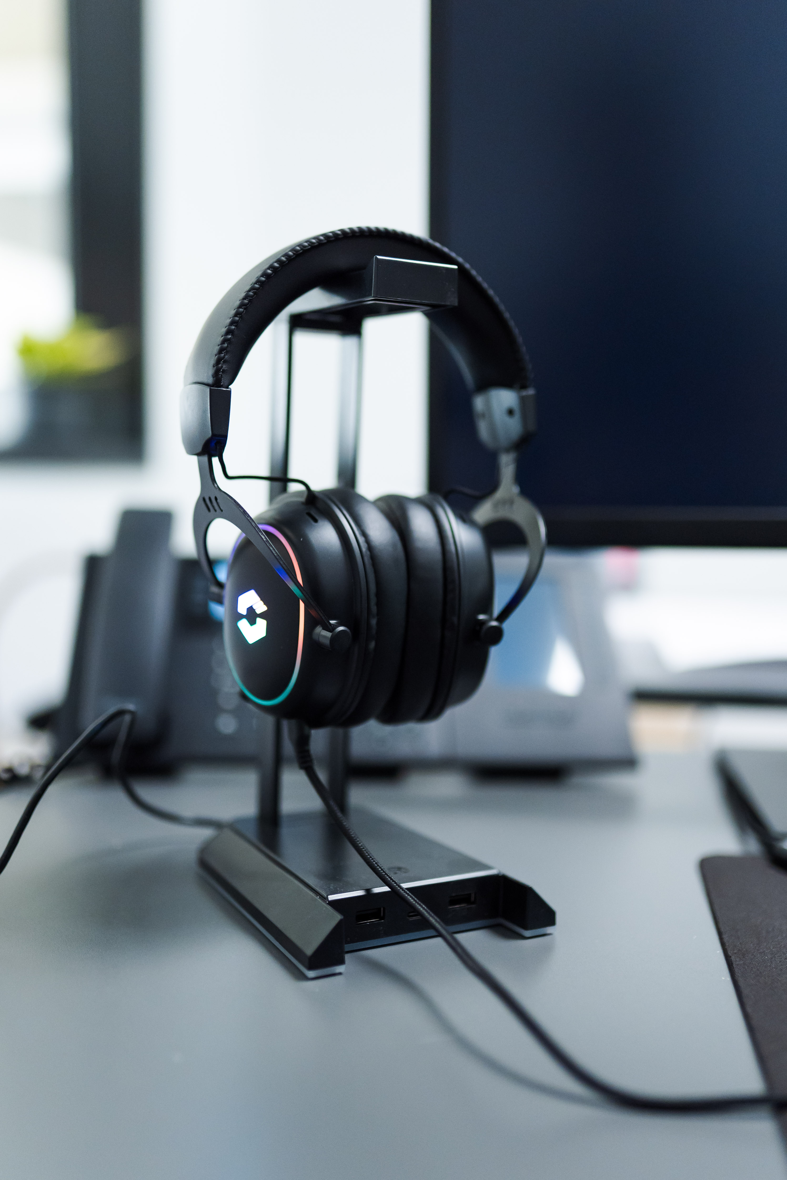 Schwarzer Gaming-Kopfhörer mit bunten LED-Lichtern steht auf einem Headset-Ständer auf einem modernen Schreibtisch neben Monitor und Telefon