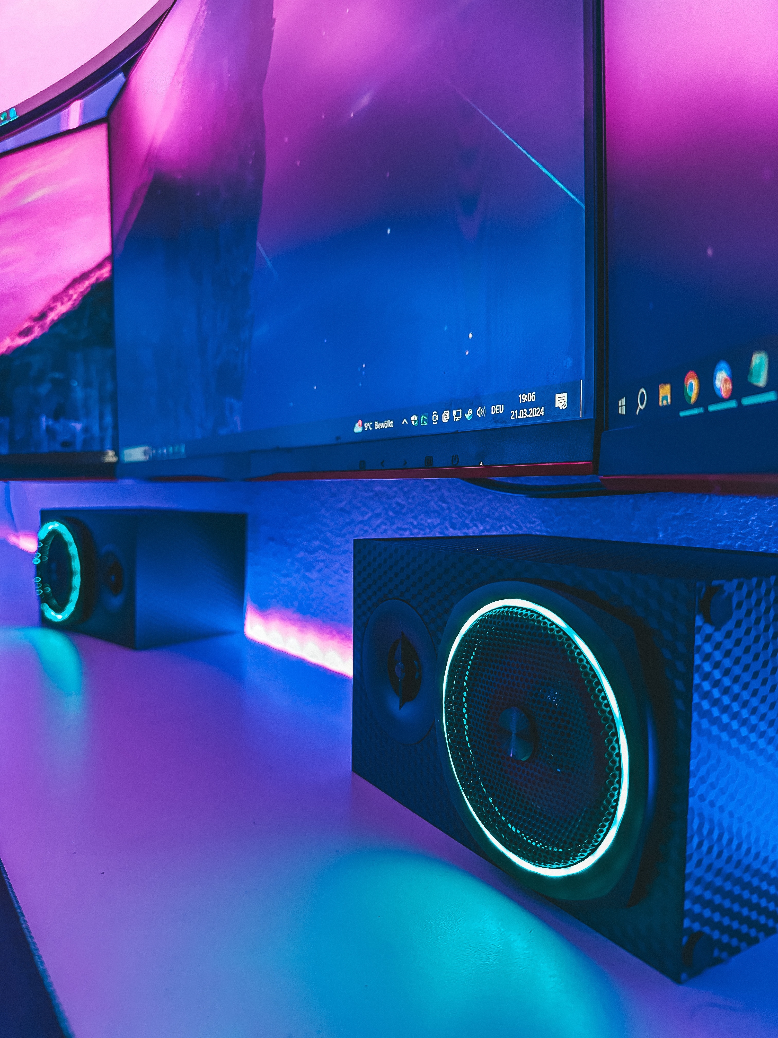 Seitenansicht eines modernen Gaming-Schreibtischs mit gebogenem Monitor, LED-Beleuchtung und leuchtenden Lautsprechern in Blau und Pink.