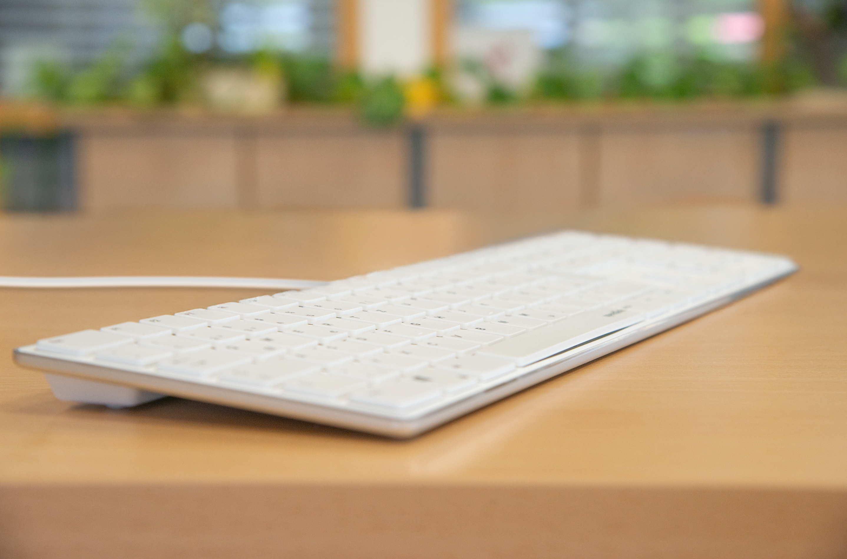 Weiße, elegante Computertastatur mit Kabel liegt auf einem Holztisch, unscharfer Bürohintergrund mit Pflanzen