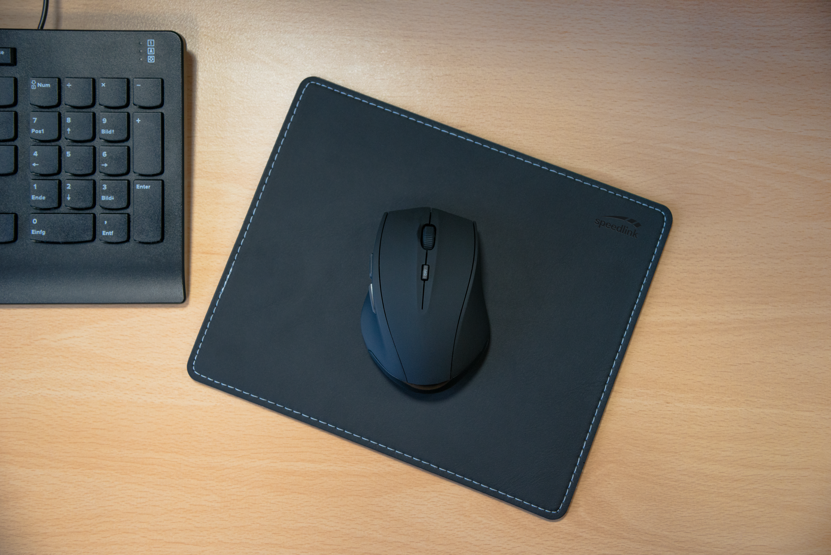 Schwarze Computermaus auf einem schwarzen Mousepad der Marke Speedlink, daneben ein Teil einer schwarzen Tastatur auf hellem Holzschreibtisch