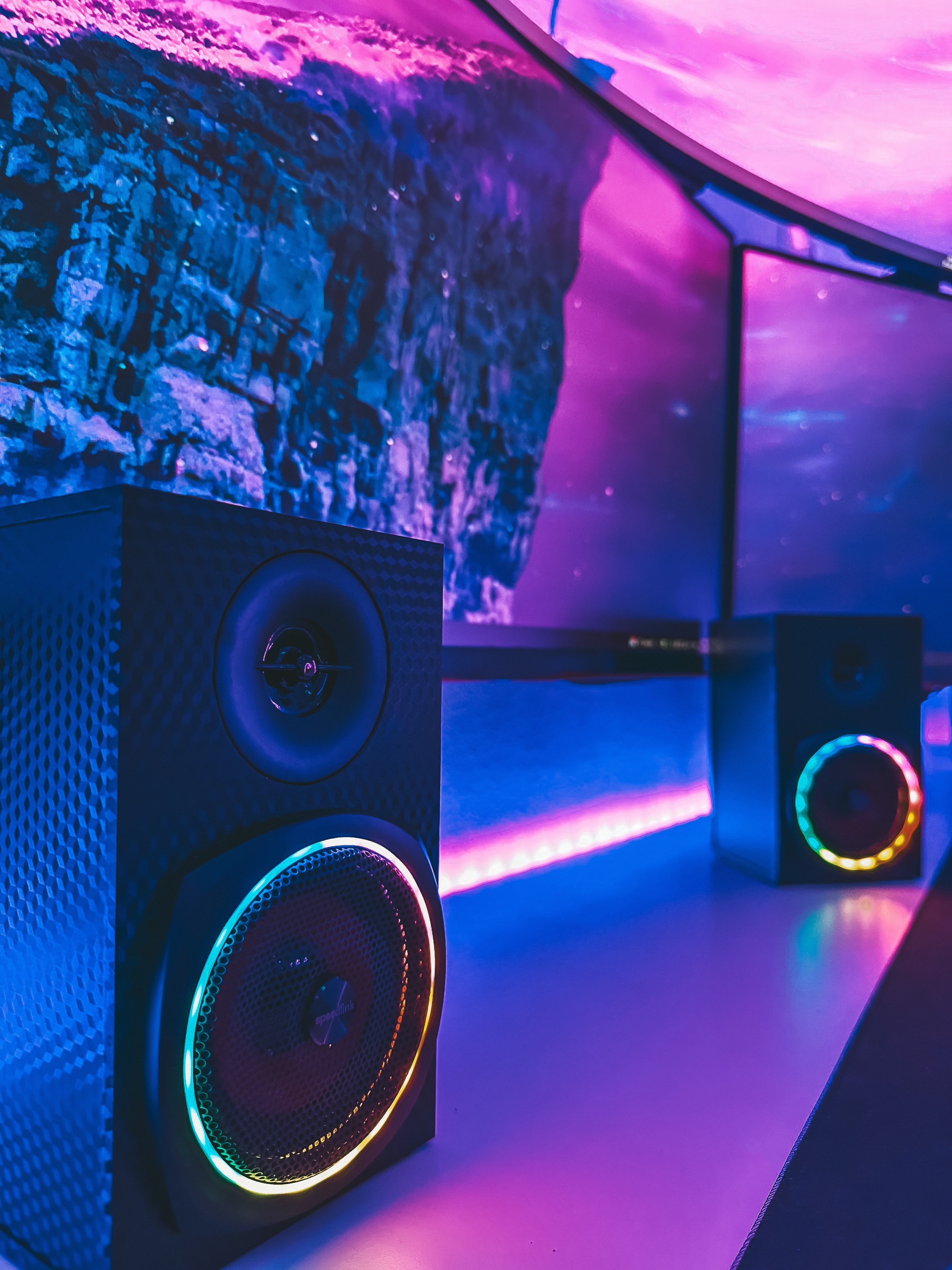 Zwei Lautsprecher mit farbiger LED-Beleuchtung auf einem Schreibtisch vor einem gebogenen Monitor mit futuristischer Grafik