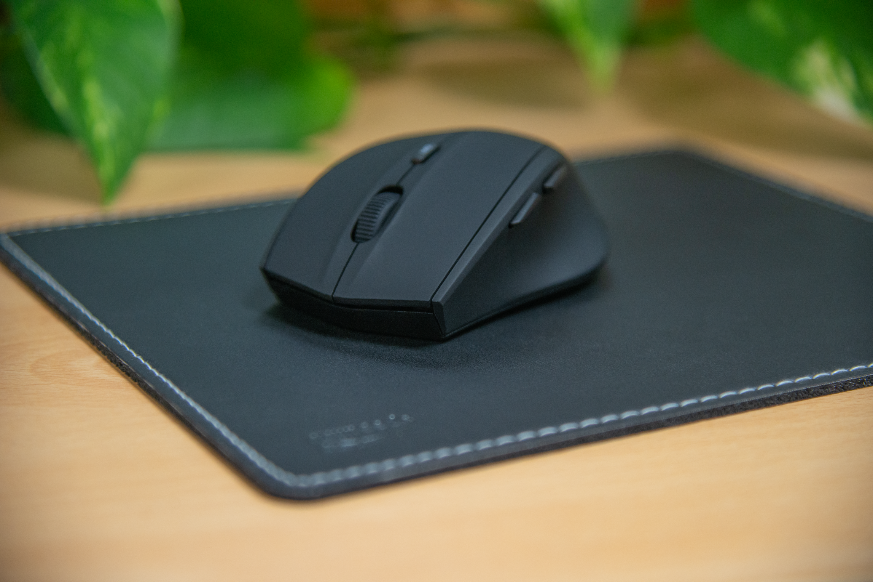 Schwarze kabellose Computermaus auf schwarzem Leder-Mauspad auf einem Holztisch, im Hintergrund grüne Pflanzen