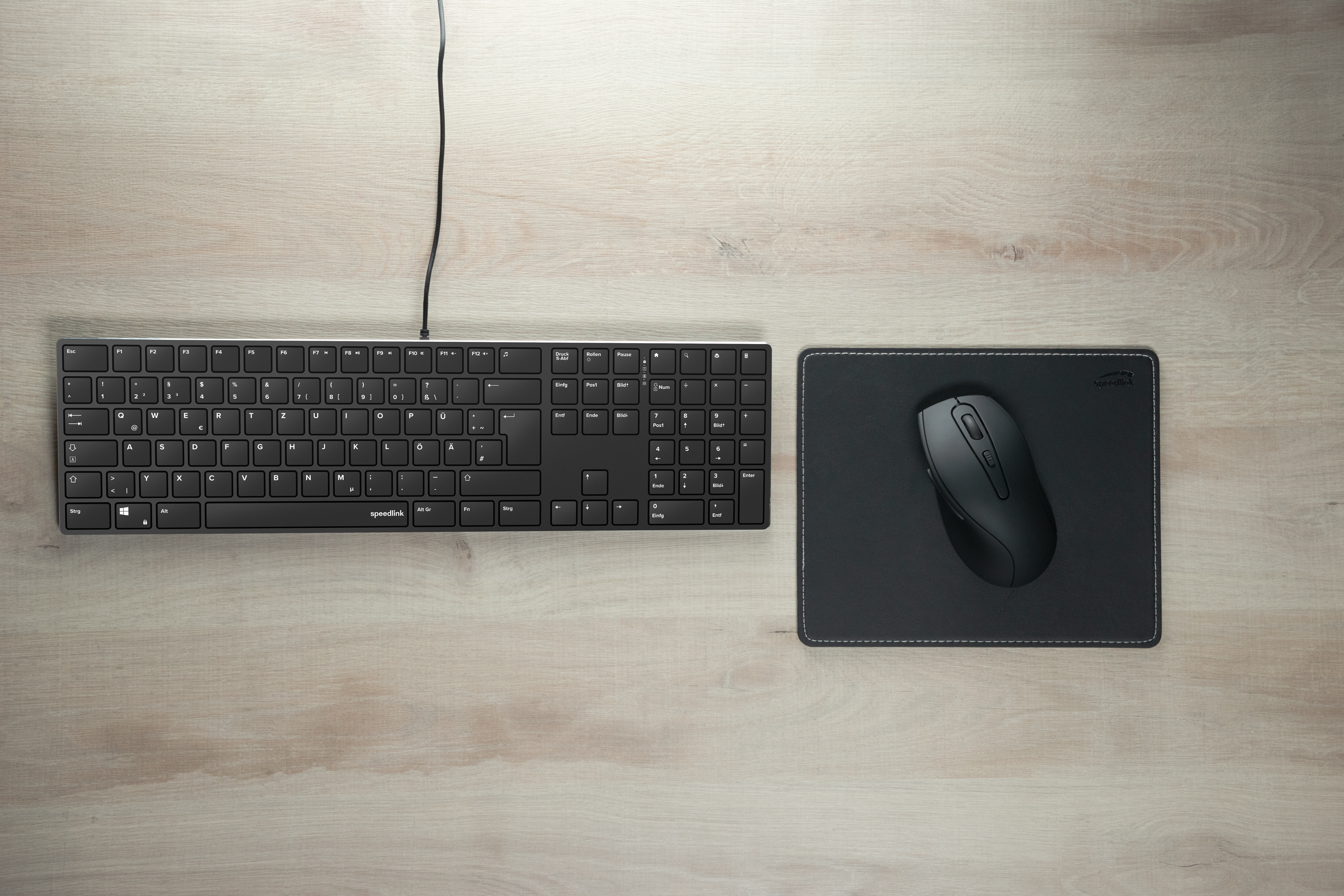 Flache schwarze Tastatur mit Kabel neben einer schwarzen kabellosen Maus auf einem Mauspad auf einem hellen Holzschreibtisch