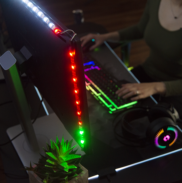 Person sitzt an einem modernen Gaming-Setup mit RGB-beleuchteter Tastatur, LED-Leiste am Monitor, Kopfhörern und einer kleinen Pflanze auf dem Schreibtisch