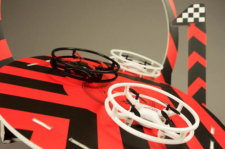 Zwei kleine Drohnen, eine schwarz und eine weiß, stehen auf einer dekorativen Rennstrecke mit roten und schwarzen Streifen und einer Zielflagge