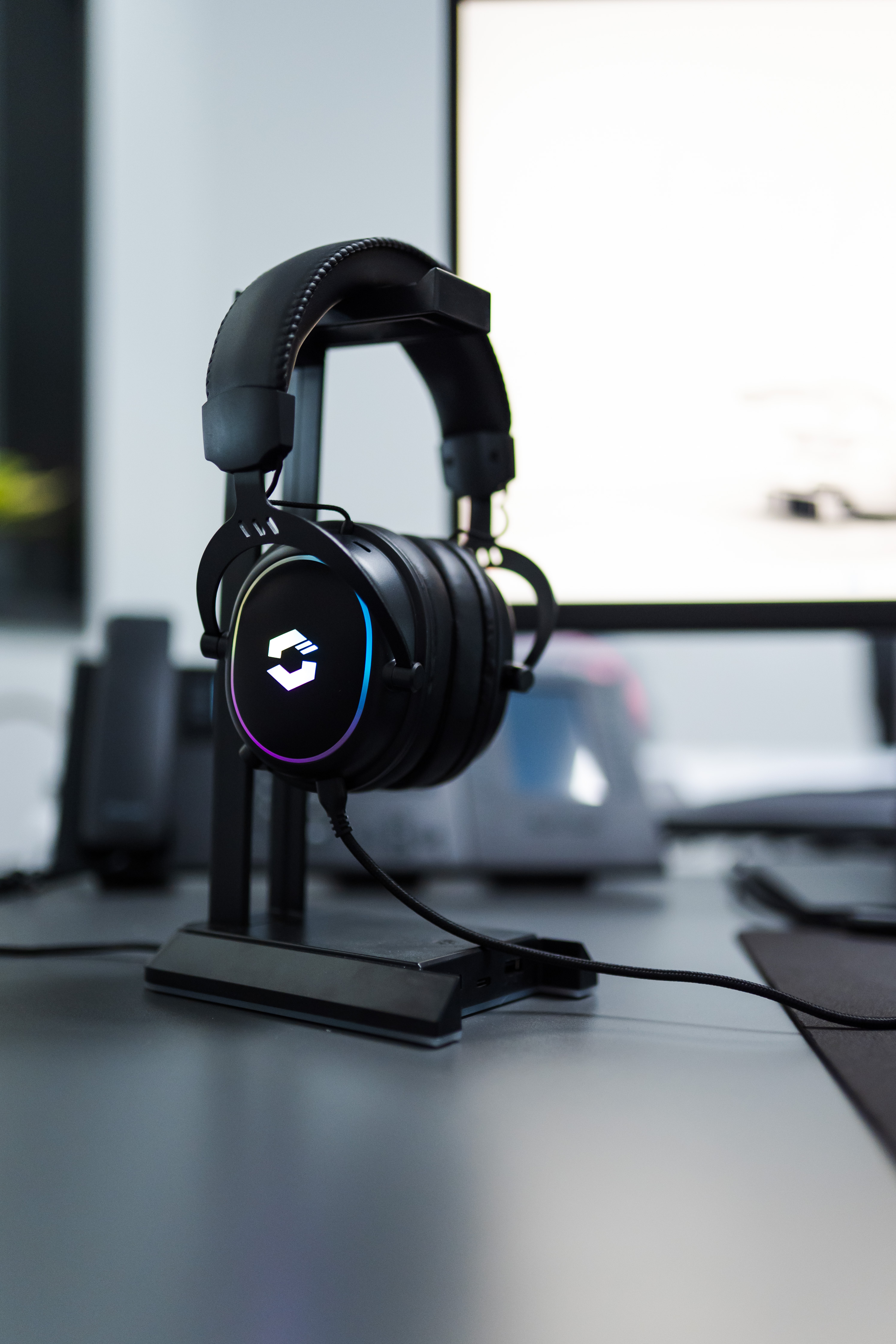 Schwarzes Gaming-Headset mit leuchtendem Logo steht auf einem Schreibtisch auf einem Kopfhörerständer vor einem Monitor